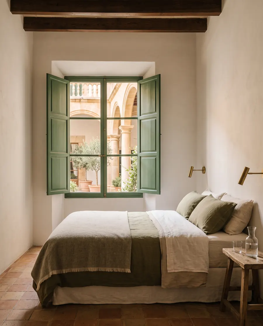A bed with sand and green linen under a tall window with green shutters open onto the courtyard arches