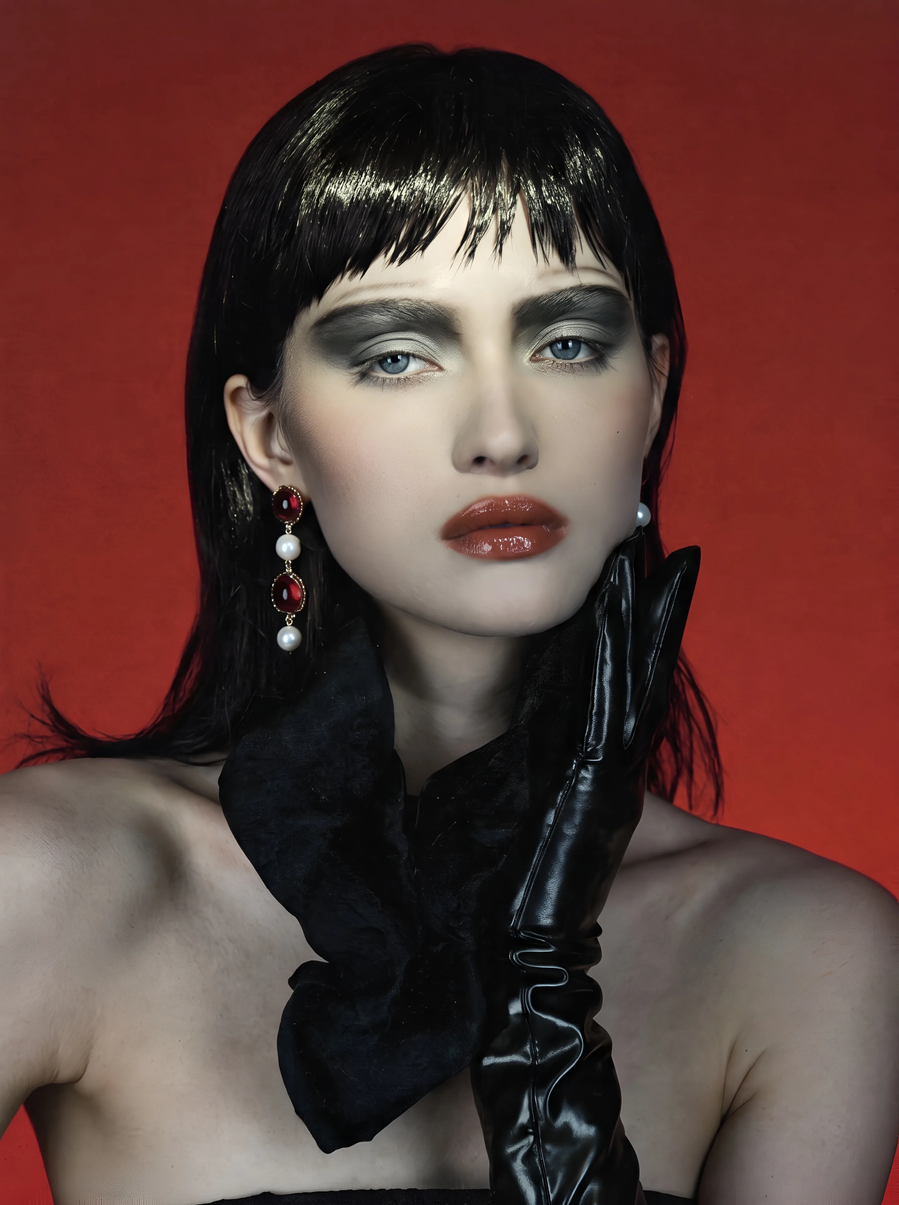 Close portrait of a woman with glossy black hair and a sharp fringe against a red wall, with smoky grey eye make-up and deep red glossy lipstick, a black velvet bow at her neck, a black leather glove at her jaw and drop earrings of red stones and pearls