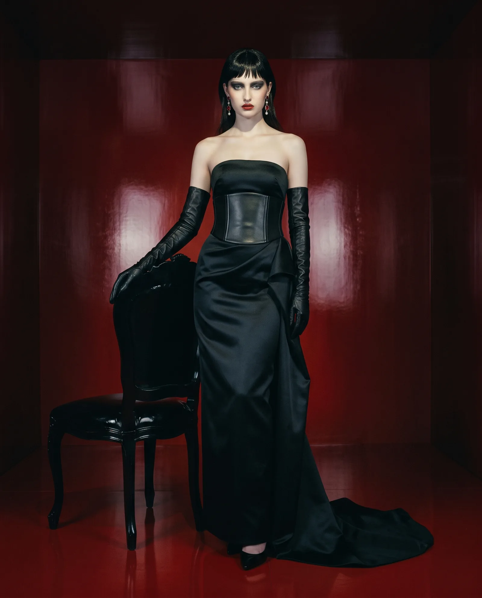 A woman with glossy black hair and a sharp fringe stands in a room of red lacquer, one hand on a black lacquer chair, in a strapless black satin gown with a wide black leather corset belt and long black leather gloves