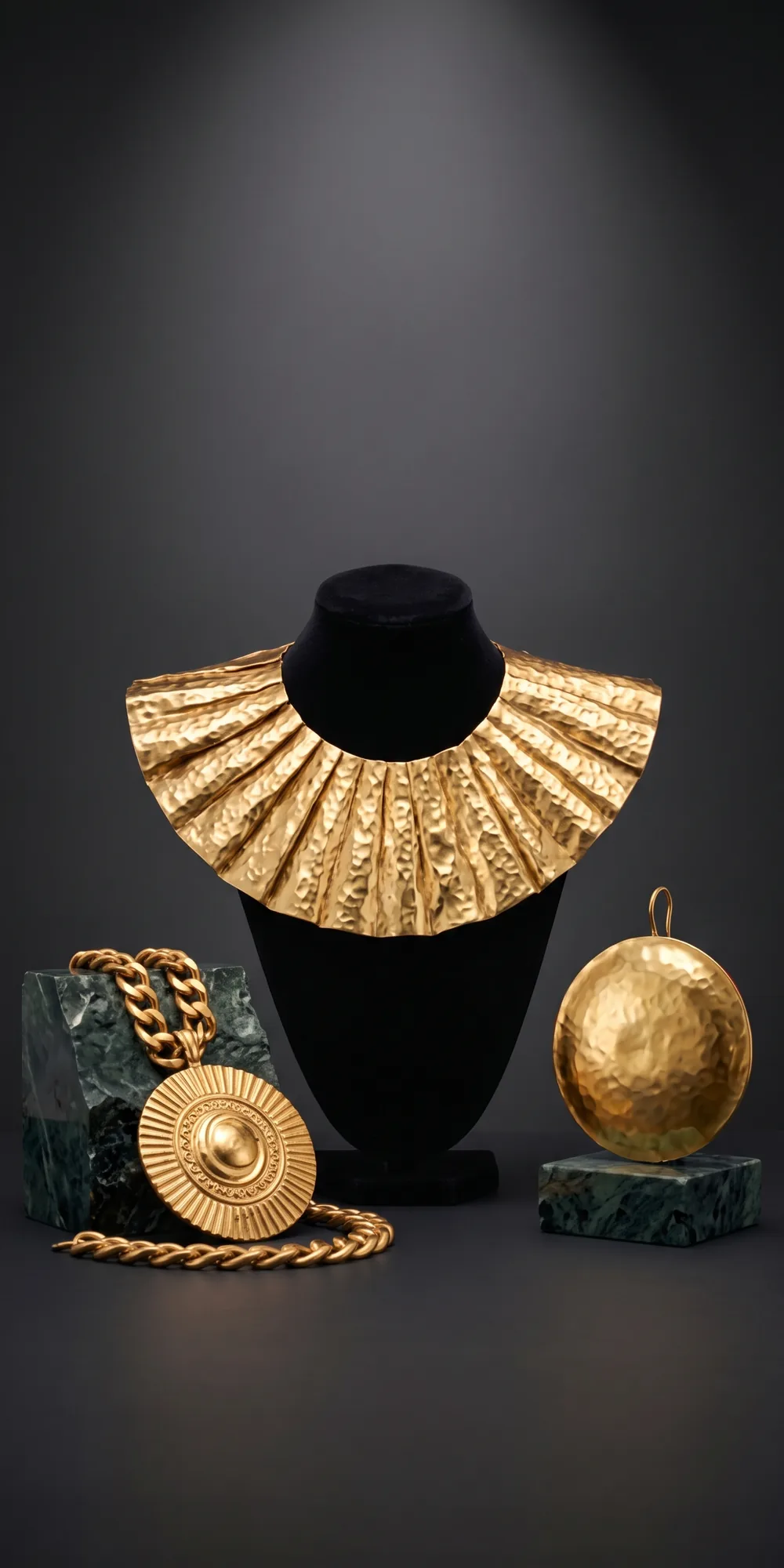 The three gold pieces: a wide hammered gold collar on a black bust, a sun disc medallion on a thick chain and a round ear disc, on dark green stone