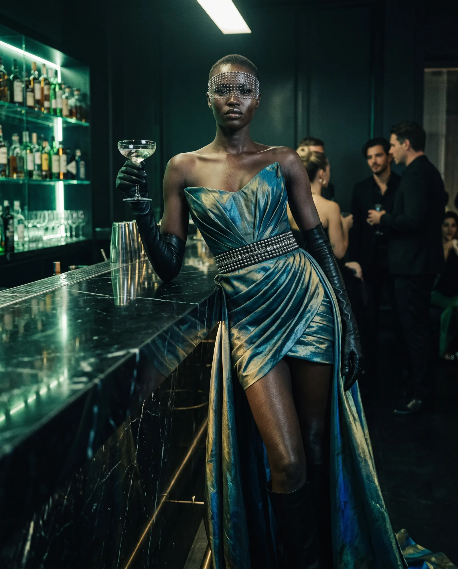 A woman with a shaved head and a crystal veil leans on the black marble counter of a cocktail bar in a strapless iridescent silk dress with a studded belt, long black gloves and a train