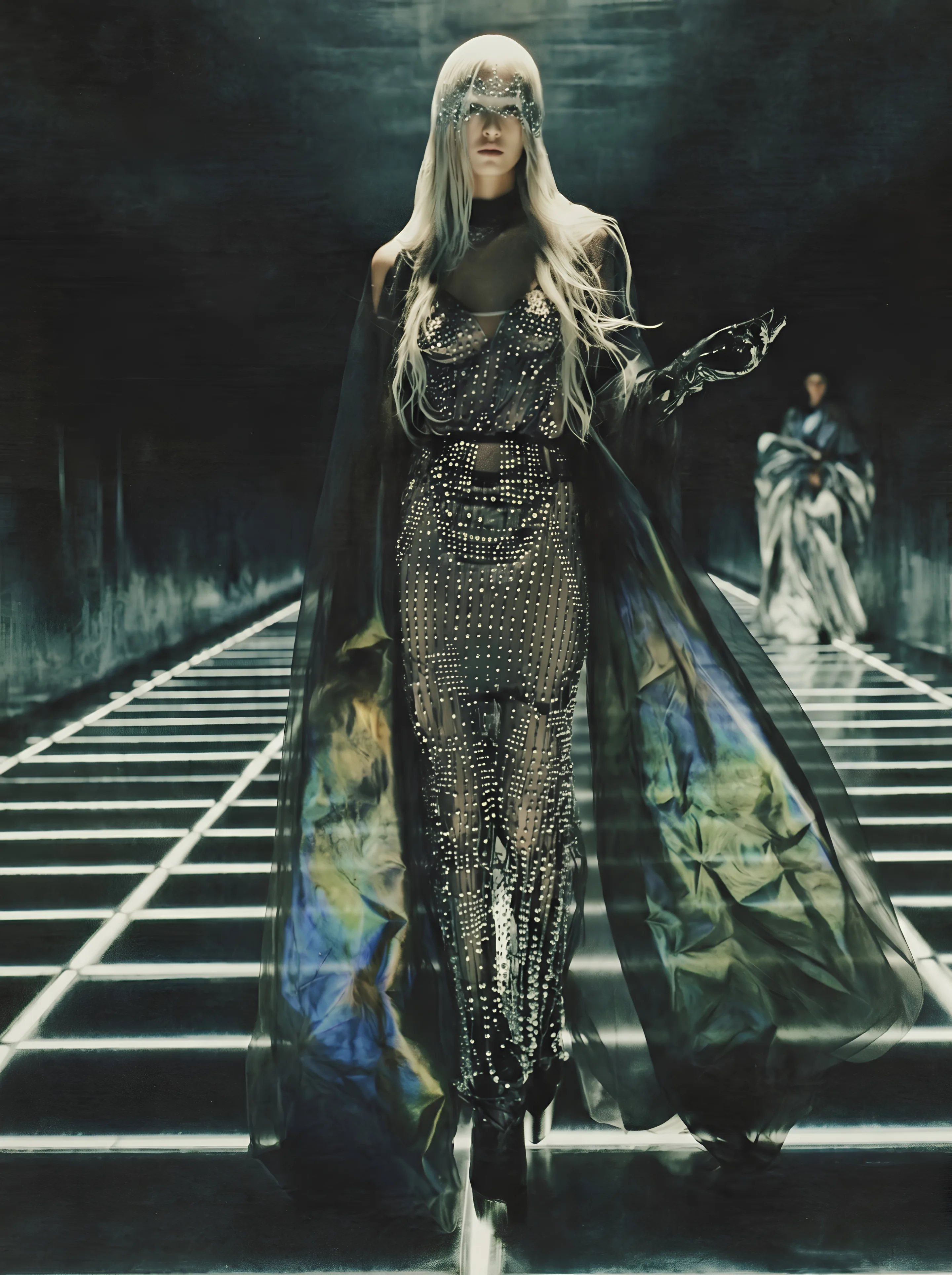 The Circuit Gown worn: a woman with long pale blonde hair walks down a dark corridor with a floor of light lines, the studded gown under the sheer cape with an iridescent lining, a crystal veil over her eyes