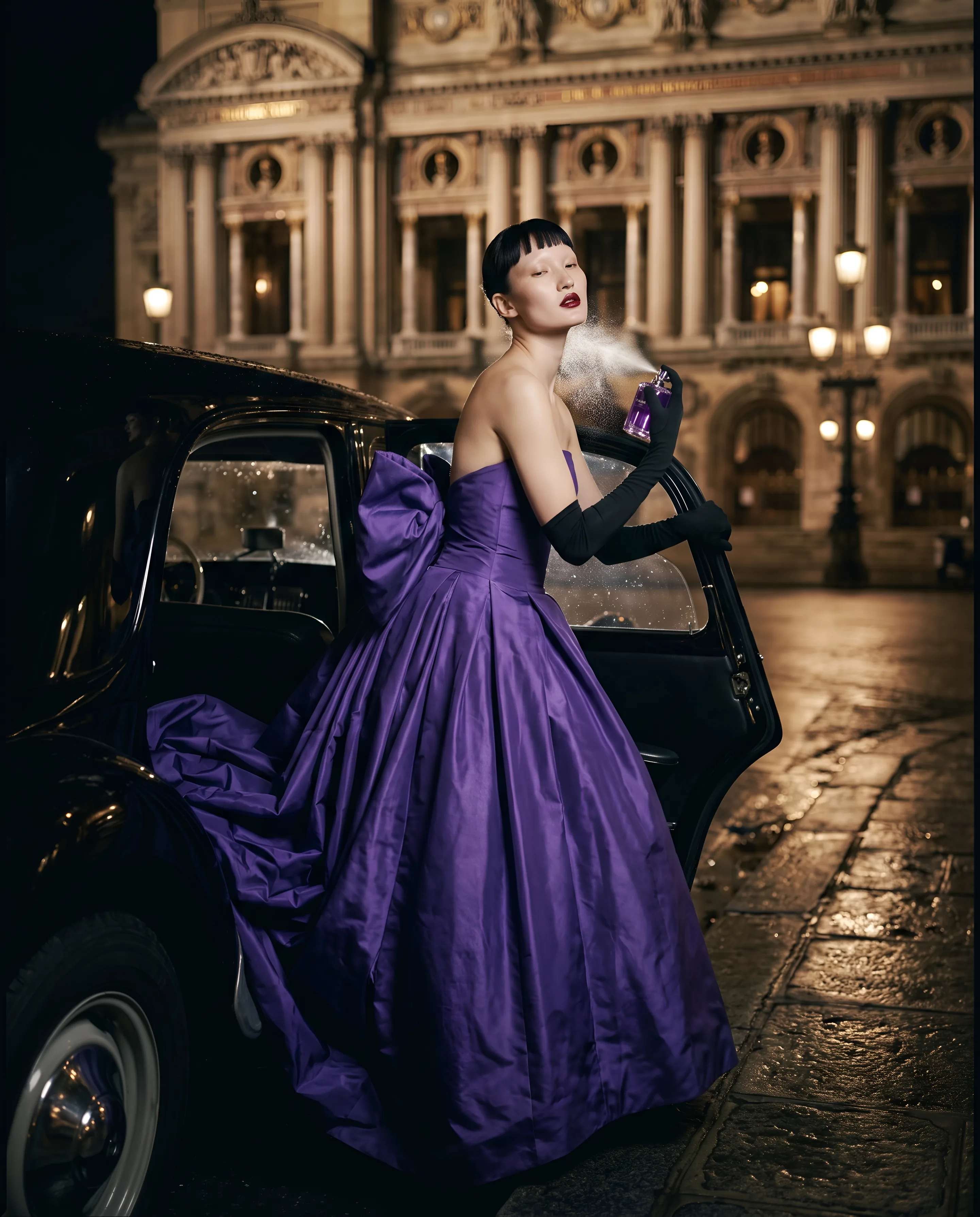A woman with a short black fringe stands at the open door of a black car in front of an opera house at night, in a violet taffeta gown, spraying perfume on her neck