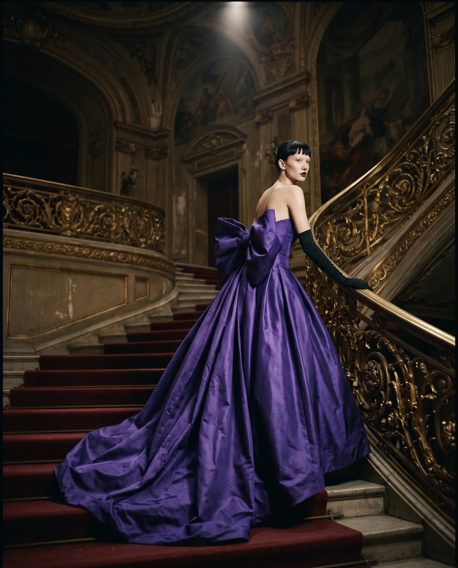 A woman with a short black fringe climbs the red carpet of an opera staircase in a strapless violet taffeta gown with a huge bow at the back and long black gloves
