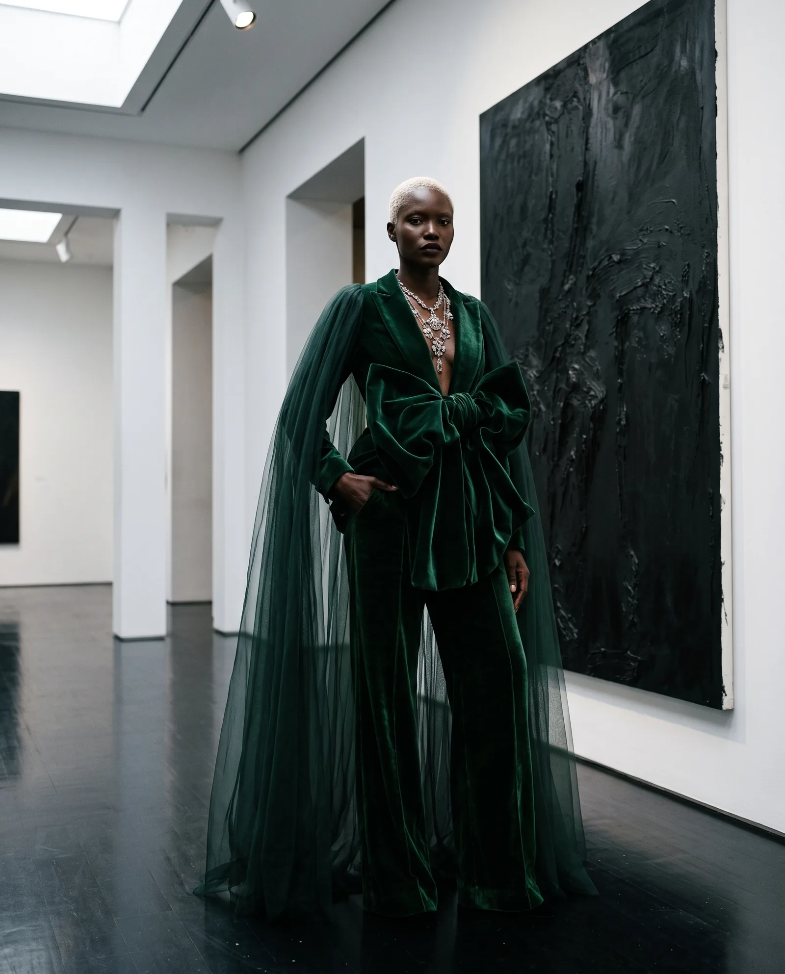 A model with cropped bleached hair stands in a white gallery beside a large black painting, in a bottle green velvet suit with a bow at the waist, a tulle cape and a layered silver necklace