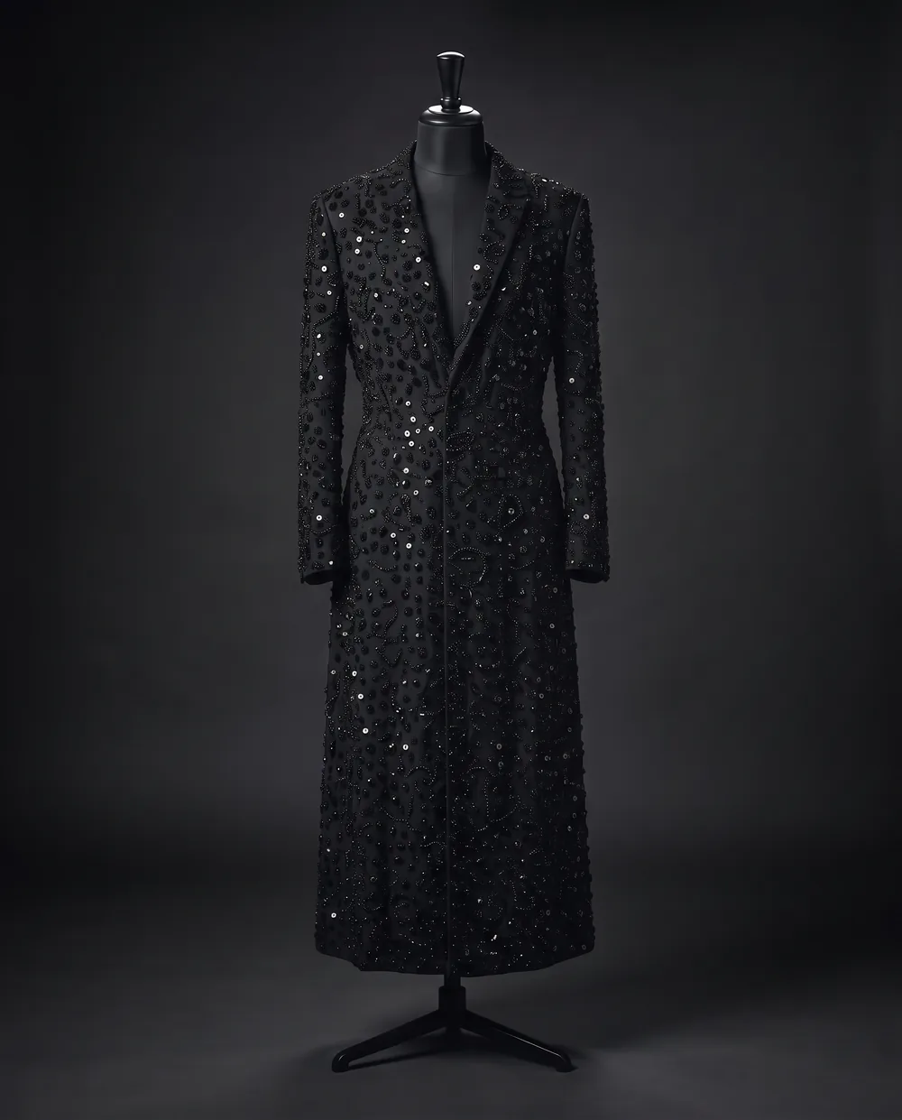 The Beaded Coat on a black dress form: a long coat covered in black glass beads from collar to hem