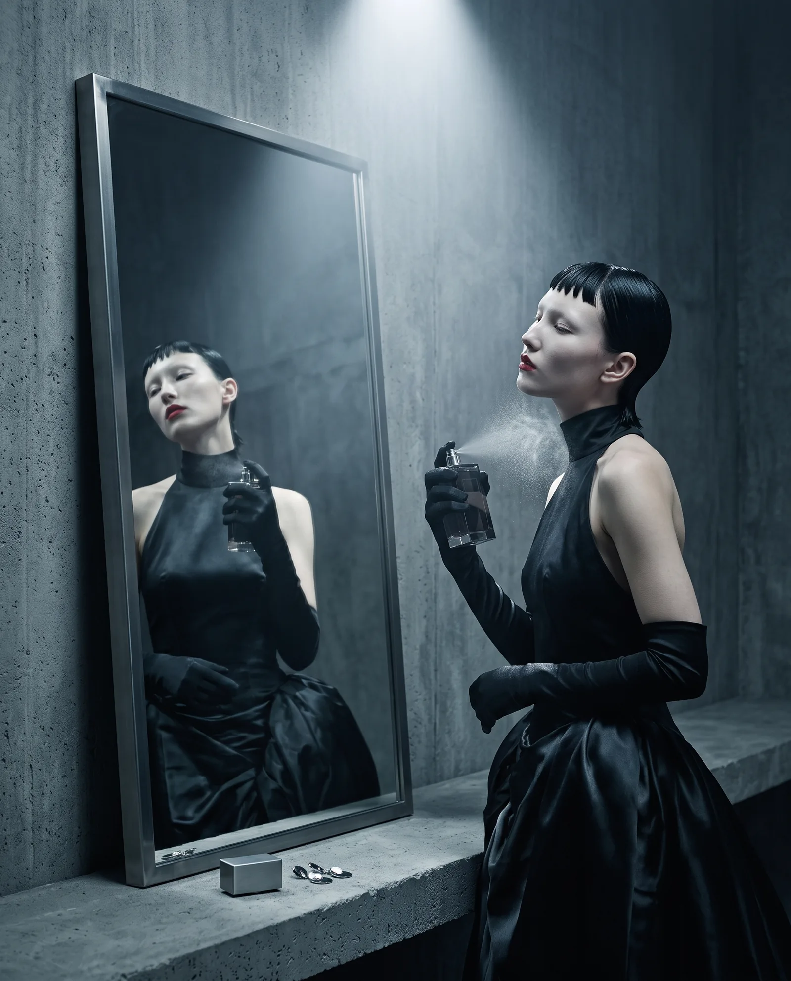 A woman with a short black fringe sprays perfume on her neck in front of a steel framed mirror, in a black satin gown and long gloves